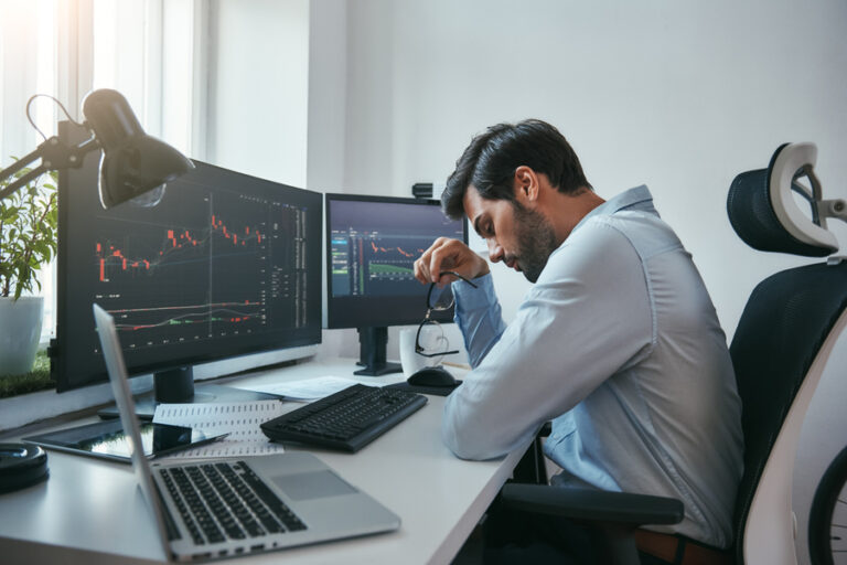 Time to take a break. Side view of tired young Forex trader holding eyeglasses and feeling tired while working with data and charts on computers at his modern office. Forex market.