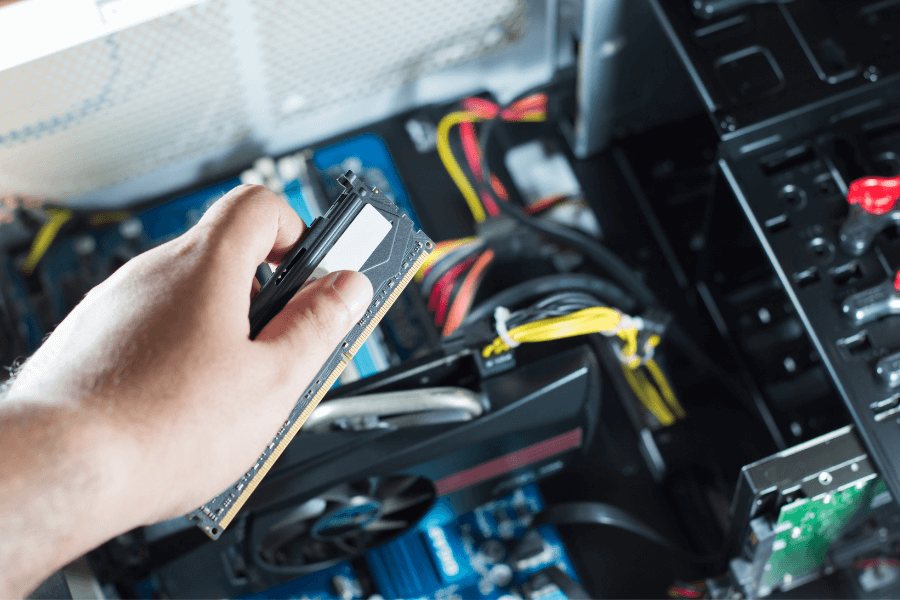 a hand holding the RAM of a desktop computers and in the background the internal systems of the PC in show